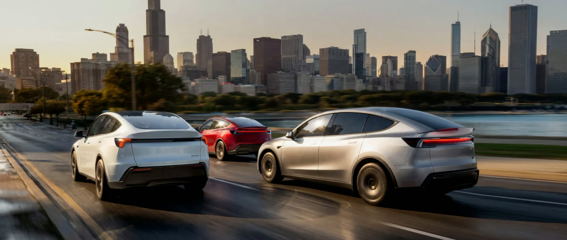 Three Tesla Model Y vehicles driving along a waterfront road at sunset